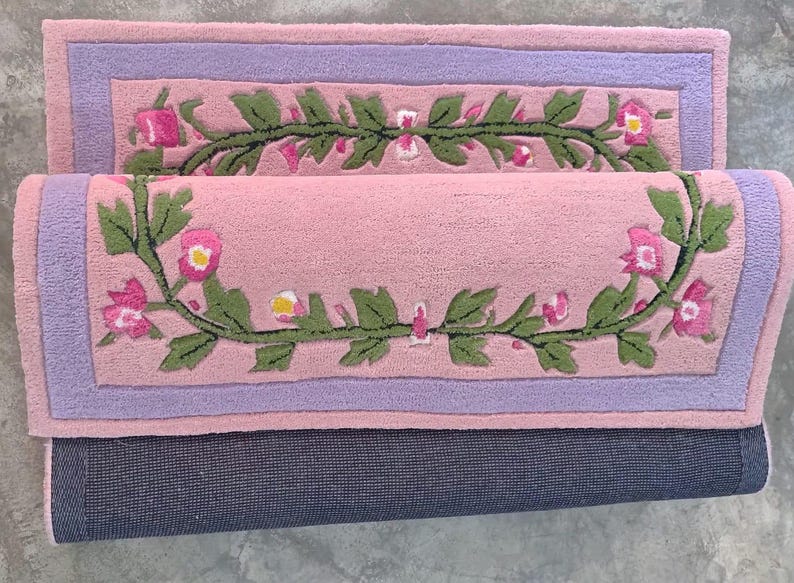 May include: A rectangular tufted rug featuring a soft pink centre with a green vine and pink flower motif, framed by a lavender border. The rug is folded over to reveal a dark grey textured backing on the underside of the fabric material.