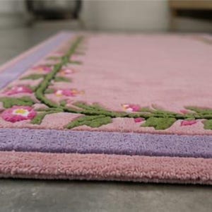 May include: A rectangular rug featuring a soft pink centre field bordered by a lavender band and a green vine motif with pink flowers. The tufted texture creates a raised surface pattern, with the floral design framing the perimeter of the rug.