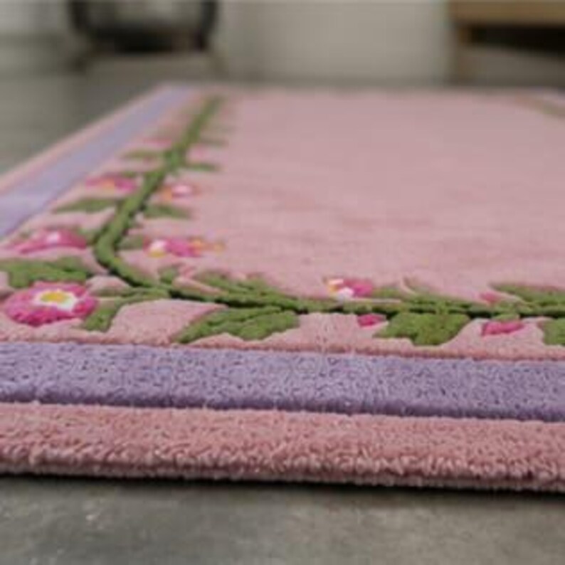 May include: A rectangular rug featuring a soft pink centre field bordered by a lavender band and a green vine motif with pink flowers. The tufted texture creates a raised surface pattern, with the floral design framing the perimeter of the rug.