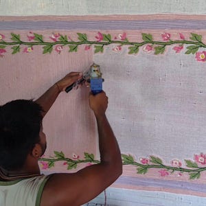 May include: A rectangular rug on a frame featuring a light pink centre and a decorative border of green vines with pink flowers. A blue tufting gun is being used to fill in the rug surface, which is partially covered in light pink yarn.