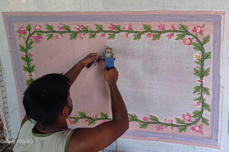 May include: A rectangular rug on a frame featuring a light pink centre and a decorative border of green vines with pink flowers. A blue tufting gun is being used to fill in the rug surface, which is partially covered in light pink yarn.