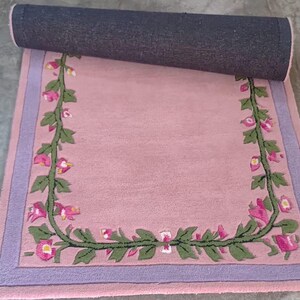 May include: A rectangular area rug featuring a soft pink centre bordered by a lavender frame. A continuous green vine with pink and white flowers forms a U-shaped pattern along the inner edges of the rug, which is partially rolled at the top.