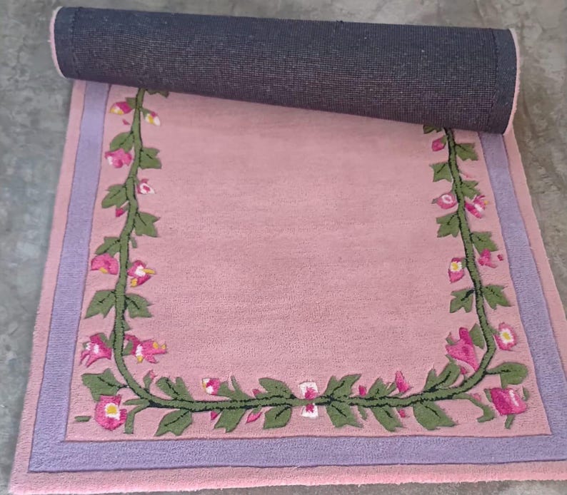 May include: A rectangular area rug featuring a soft pink centre bordered by a lavender frame. A continuous green vine with pink and white flowers forms a U-shaped pattern along the inner edges of the rug, which is partially rolled at the top.