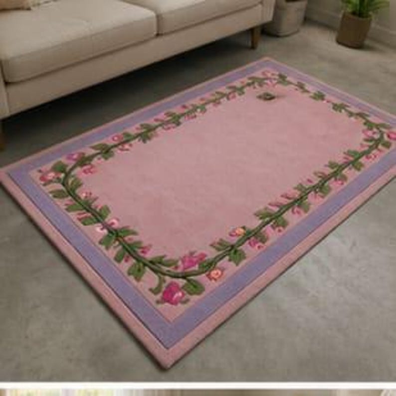 May include: A rectangular area rug featuring a solid light pink centre bordered by a decorative vine pattern with green leaves and small pink flowers. The rug is framed by a lavender border and finished with a light pink outer edge trim.