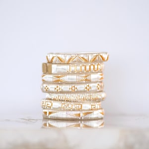 May include: A stack of seven silver bangles with gold geometric patterns. The bangles are stacked on top of each other, showcasing the intricate designs. The background is a soft, neutral color, highlighting the jewelry.