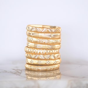 May include: A stack of eight gold-colored bangles with intricate geometric patterns. The bangles are stacked on top of each other, showcasing the detailed designs. The bangles are made of a shiny, reflective material, and the background is a soft, neutral color.