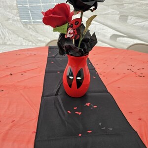 May include: A red vase featuring a Deadpool design holds a red and black rose arrangement. The vase sits on a black runner over a red tablecloth, with scattered red confetti.