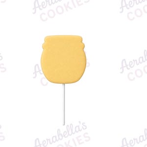 May include: A yellow honey pot shaped biscuit on a white stick. The biscuit has a smooth, flat surface and a rounded silhouette with a slightly wider top section. The background features a repeating watermark text that reads Aerabella's COOKIES.