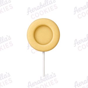 May include: A round, light beige cookie shaped like a lollipop, featuring a smooth, flat surface with a raised circular border and a centered indentation. The cookie is attached to a thin, straight, white stick extending from the bottom edge.