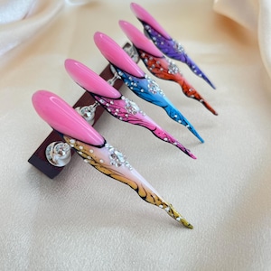Könnte beinhalten: Fünf lange, mandelförmige Kunstnägel. Jeder Nagel hat eine leuchtend rosa Spitze und ein Farbverlaufsdesign in Farben wie Blau, Orange und Lila. Verziert mit Strasssteinen, werden die Nägel auf einem dunklen Holzständer präsentiert.
