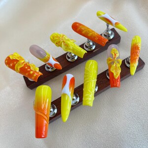 Könnte beinhalten: Eine Sammlung von langen Acrylnägeln in leuchtenden Farben. Die Nägel zeigen Farbverläufe von Gelb, Orange und Weiß, einige sind mit floralen und abstrakten Mustern verziert. Jeder Nagel wird auf einem silbernen Ständer vor einem neutralen Hintergrund präsentiert.