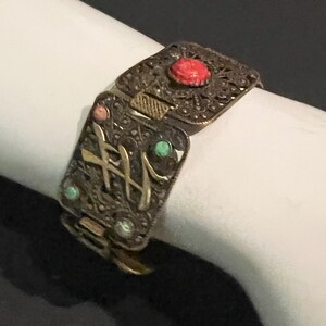 以下が含まれることがあります： 繊細なフィリグリー模様が施された装飾的な金属製ブレスレット。中央に赤い石、小さなターコイズとコーラルのアクセントが特徴です。ヒンジ付きのデザインと留め金があり、アンティークまたはヴィンテージの美学を示しています。