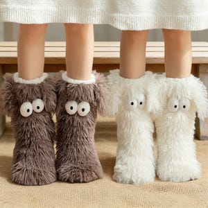 Puede incluir: Dos pares de zapatillas novedosas, una marrón y otra blanca. Las zapatillas tienen forma de perros peludos, con grandes ojos saltones y pelaje esponjoso. Cada zapatilla tiene un ribete blanco alrededor del tobillo.