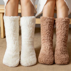 May include: Two pairs of cozy, knee-high socks. One pair is a soft cream color, and the other is a warm brown. The socks have a fluffy, textured appearance, suggesting a plush, comfortable material. Perfect for lounging or keeping warm.