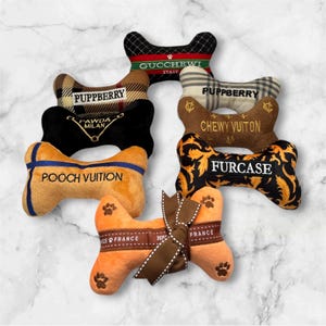 May include: A selection of dog bone-shaped toys in different colours and designs. The toys display text including "POOCH VUITION", "FURCASE", and "GUCCHEWI ITALY". Some bones have paw prints and ribbon details.