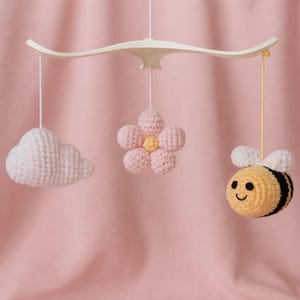 May include: A handmade crochet baby mobile featuring a white cloud, a pink flower, and a yellow bee with black stripes and white wings. The mobile is suspended from a cream-colored curved bar against a pink backdrop.
