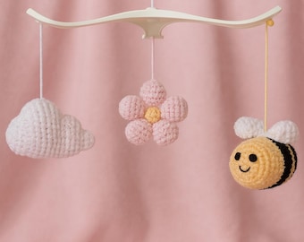 May include: A handmade crochet baby mobile featuring a white cloud, a pink flower, and a yellow bee with black stripes and white wings. The mobile is suspended from a cream-colored curved bar against a pink backdrop.