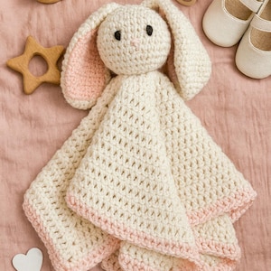 Op de afbeelding: Een crème gehaakte konijn knuffel met roze accenten, omringd door babyaccessoires. Inclusief een houten ster, een fopspeen, een borstel en witte babyschoenen. De knuffel heeft een zachte, getextureerde uitstraling.