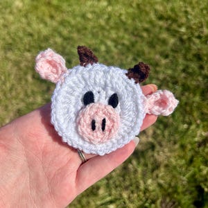 Puede incluir: Una cara de vaca hecha a mano con ganchillo, con una base blanca, orejas y hocico rosas, ojos negros y cuernos marrones. El objeto redondo tiene una textura, probablemente de hilo. Se sostiene en una mano sobre un fondo verde borroso.