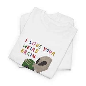 May include: A white t-shirt featuring a graphic print with the text I LOVE YOUR WEIRD BRAIN in multicolored letters. Below the text is an illustration of a grey alien figure holding a green circular object with a dark center.