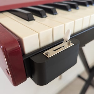 May include: Close-up of a red and black keyboard instrument with ivory and black keys. A white switch is mounted on the side. The instrument is on a black stand.