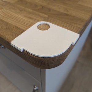 May include: A white corner protector with a circular handle hole is placed on a wooden countertop. The countertop has a light brown wood grain pattern. The corner protector is designed to fit the rounded edge of the countertop.