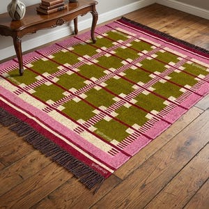 Op de afbeelding: Een rechthoekig vloerkleed met een geometrisch rasterpatroon in olijfgroen, crème en donkerrood. Het kleed is omrand met brede roze en donkerrode banden en afgewerkt met donkerbruine franjes aan de korte zijden. De tekst gudiyarugs is zichtbaar.
