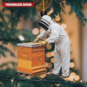 May include: Ornament featuring a beekeeper in a white suit and protective headgear tending to a wooden beehive. The beehive is orange and yellow with the text "YOUR NAME". The ornament has the text "PERSONALIZED DESIGN".