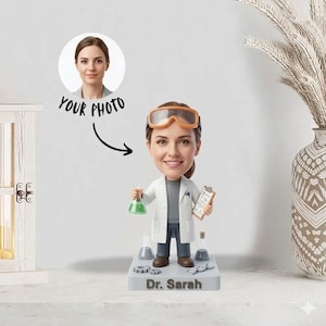 Può includere: Una statuetta bobblehead di una scienziata con camice bianco e occhiali protettivi, che tiene in mano un pallone con liquido verde e un blocco per appunti. La base grigia ha il nome "Dr. Sarah". Una foto è mostrata con il testo "YOUR PHOTO".