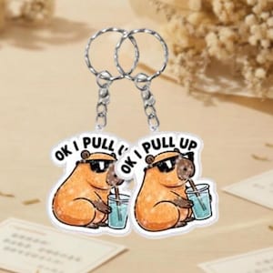 May include: Two keychains featuring a cartoon capybara wearing sunglasses and drinking from a cup. The text "OK I PULL UP" is printed on each keychain. The keychains have silver metal rings.