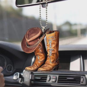 May include: A brown cowboy hat and boots car charm hanging from a silver beaded chain. The boots have the words "Name here" in white script. The charm is suspended in front of a car's dashboard.