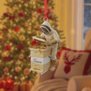 May include: A Christmas ornament featuring a beekeeper inspecting a wooden beehive. The ornament is cream-colored with a red ribbon for hanging. The beehive has the name "Jessica" on it. The background includes a Christmas tree and a decorative pillow.