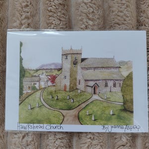 May include: A watercolor print of Hawkshead Church, featuring a stone church with a clock tower, set in a green landscape with a winding path and gravestones. The artwork is signed by Joanna Aspap.