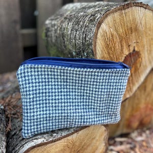 May include: A small, rectangular pouch with a blue and white houndstooth pattern. The pouch has a blue zipper and is made of a textured fabric. The background features a wooden log and a wooden fence.