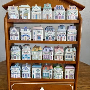 Op de afbeelding: Een houten kruidenrek met een lade, met 20 keramische kruidenpotjes in de vorm van miniatuurhuisjes. Elk potje is voorzien van een kruidennaam en heeft kleurrijke details. Het rek is gemaakt van donker hout en heeft een decoratieve bovenkant.