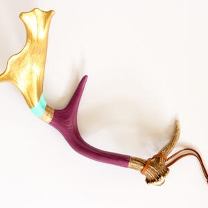 Könnte beinhalten: Ein dekoratives Geweih, bemalt in Gold, Lila und Türkis. Das Geweih hat einen Lederriemen und ist vor einem weißen Hintergrund platziert. Der goldene Abschnitt befindet sich oben, mit türkisfarbenen und goldenen Streifen, die in Lila übergehen.