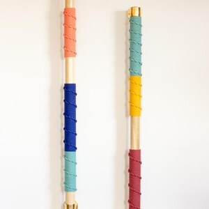 May include: Two wooden wall-mounted decorative poles. Each pole is wrapped with colorful rope in sections of coral, blue, and teal, and teal, yellow, and burgundy. The poles have gold-colored metal hardware at the top and bottom. The background is a plain white wall.