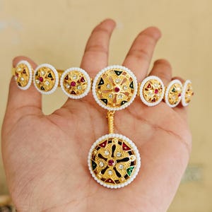May include: A gold hand chain with a floral design. The chain has multiple circular elements with white borders, gold filigree, and red, green, and clear accents. A larger pendant hangs below, also with a floral design.