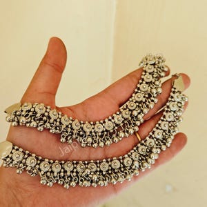 May include: Two silver anklets with detailed floral and beaded patterns. Each anklet has small, round beads and floral-shaped decorations. The anklets are shown on a hand, highlighting their delicate craftsmanship and intricate design.