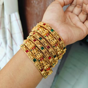 May include: Ornate gold bangles featuring detailed floral patterns and set with red and green gemstones. The bangles are stacked on a wrist, displaying their intricate design and vibrant colours.