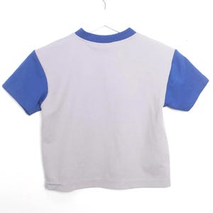 Puede incluir: Una camiseta de manga corta con cuerpo blanco y mangas y cuello azules. La camiseta est&aacute; colgada de una percha blanca sobre un fondo blanco. La camiseta es de color s&oacute;lido.