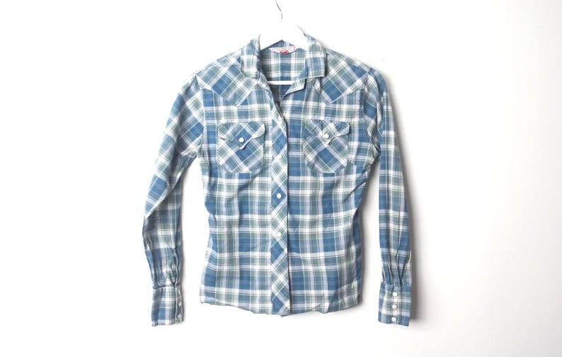 May include: A blue, green, and white plaid button-down shirt with long sleeves. The shirt has two chest pockets with button closures and a classic collar. The cuffs have button closures. The shirt is hanging on a white hanger.