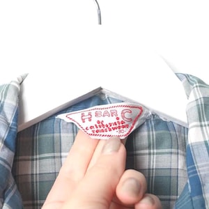 May include: Close-up of a blue, white, and green plaid shirt. The shirt is hanging on a white hanger. A red and white label reads "H BAR C California Ranchwear".