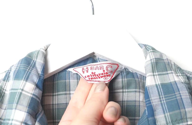May include: Close-up of a blue, white, and green plaid shirt. The shirt is hanging on a white hanger. A red and white label reads "H BAR C California Ranchwear".