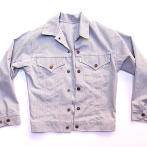 May include: A light grey denim jacket featuring a pointed collar, long sleeves with buttoned cuffs, and a front button closure. The jacket includes two chest pockets with pointed flaps and copper-coloured buttons throughout the front and cuffs.