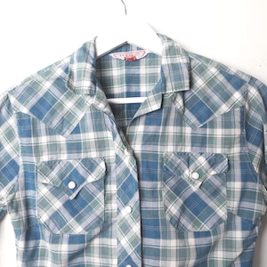May include: A blue, green, and white plaid shirt with two chest pockets and pearl snap closures. The long-sleeved shirt is hanging on a white hanger against a white background. The shirt has a classic western style.