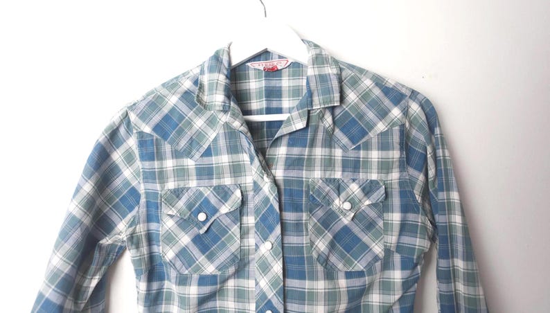 May include: A blue, green, and white plaid shirt with two chest pockets and pearl snap closures. The long-sleeved shirt is hanging on a white hanger against a white background. The shirt has a classic western style.