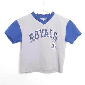 Puede incluir: Una camiseta gris y azul de manga corta con la palabra "ROYALS" arqueada en el pecho en letras azules y doradas. La camiseta tiene cuello en V y un peque&ntilde;o logotipo en la parte inferior derecha. La camiseta cuelga sobre un fondo blanco.