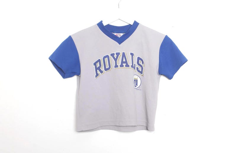 Puede incluir: Una camiseta gris y azul de manga corta con la palabra "ROYALS" arqueada en el pecho en letras azules y doradas. La camiseta tiene cuello en V y un peque&ntilde;o logotipo en la parte inferior derecha. La camiseta cuelga sobre un fondo blanco.