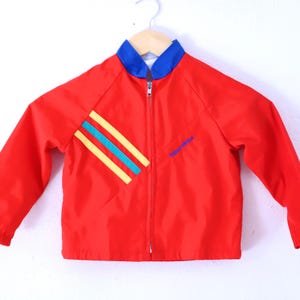 Puede incluir: Chaqueta roja para niños con cuello azul y cierre de cremallera. La chaqueta presenta un diseño diagonal con rayas amarillas y turquesas. La palabra "windbreaker" está impresa en azul en el lado derecho.